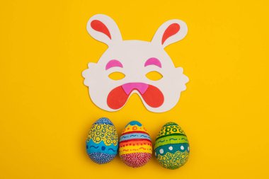 Tatlı Paskalya tavşanı maskesi ve sarı arka planda renkli yumurtalar. Paskalya dekorasyonu