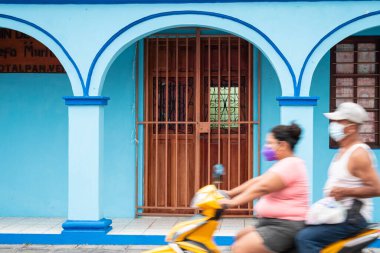 TLACOTALPAN, VERACRUZ, MEXICO - 20 AĞUSTOS 2021: Bir çift yerli motosiklet sürüyor, Tlacotalpan, Veracruz, Meksika 'da tipik bir mavi evi geçiyor