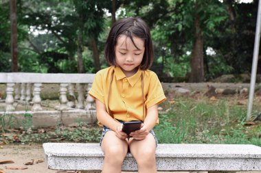 Parkta cep telefonu kullanan mutlu kız çocuğu. Çocuk ekrandaki cihazlara bakıyor. İletişim kavramı, teknoloji.