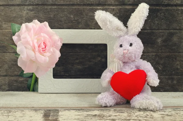 Lovely rabbit doll holding red heart shape sitting near blank fr — Stock Photo, Image