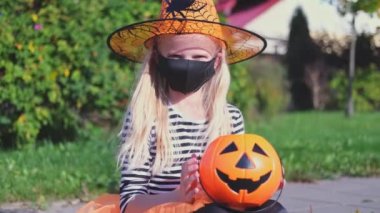 4 bin. Cadılar Bayramı çocukları maskesi. Cadı kostümlü sarışın kız ve balkabağı kovası. Çocuk dışarıda şeker yemek için siyah yüz maskesi takıyor. COVID-19 'dan koruma