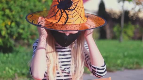 4k. Halloween kindermasker. blond meisje in heksenkostuum en oranje hoed. Kind zet buiten zwarte gezichtsmaskers op ter bescherming tegen COVID-19 — Stockvideo