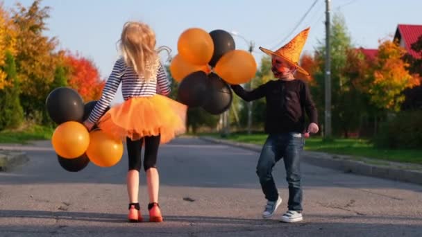 Langzame beweging. Halloween kinderen. Broer en zus in carnaval kostuums buiten. Jongen en meisje hebben plezier met oranje en zwarte ballonnen. — Stockvideo