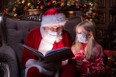 Çocuklar ve Noel Baba tıbbi maskeli. Kız ve Noel Baba evde Noel ağacının yanında oturmuş Noel kitabı okuyorlar. Mutlu yıllar, 2021.