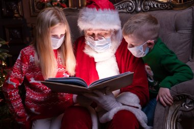 Çocuklar ve Noel Baba tıbbi maskeli. Kız, erkek ve Noel Baba evde Noel ağacının yanında oturmuş Noel kitabı okuyorlar. Mutlu yıllar, 2021.