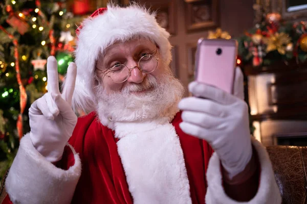 サンタからクリスマスオンラインお祝い。遠くに携帯電話を使用してサンタクロースクリスマスは人々と子供を呼び出します。自宅でクリスマスツリーの近くに座っている。新年明けましておめでとうございます｜Covid 2021 — ストック写真