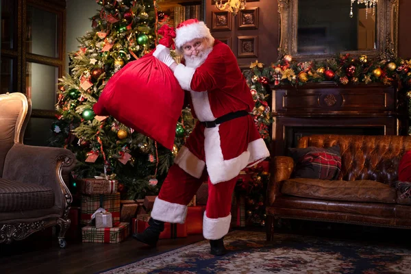 Dansande Santa förbereder sig för att gratulera barn. Glad jultomten lyssnar musik och dansar med påse med presenter hemma nära julgranen — Stockfoto