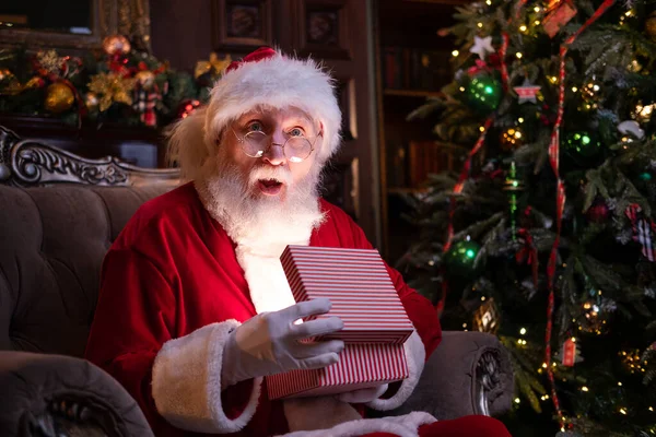 Äkta förvånad jultomten håller julklapp. Uppfyllande av önskningar. Tomten sitter hemma nära julgranen — Stockfoto