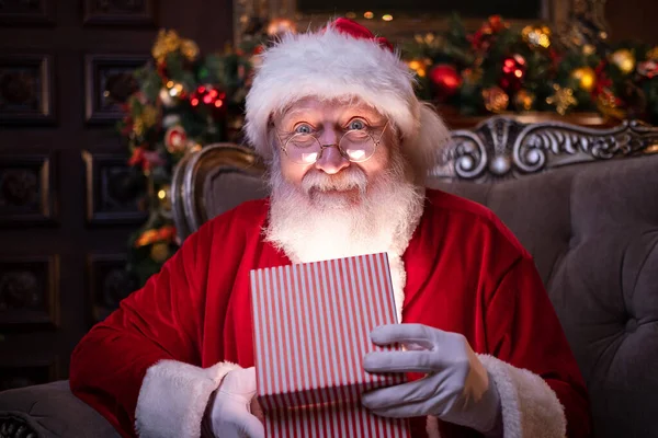 Leende jultomten håller julklapp. Uppfyllande av önskningar. Tomten sitter hemma nära julgranen — Stockfoto