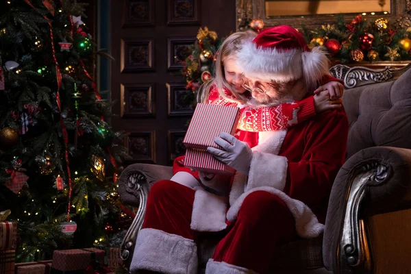 Święty Mikołaj i dziewczyna otwierając prezent świąteczny w pobliżu choinki. Dziecko przytula Mikołaja. Magiczne spełnienie pragnień — Zdjęcie stockowe