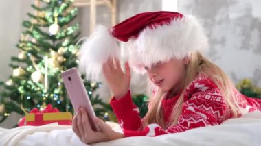 4k Christmas online tebrikler. Çocuk eğleniyor, Noel Baba şapkası ponponuyla oynuyor. Video görüşmesi için cep telefonunu kullanan gülümseyen kız. Çocuklardan bahseden arkadaşlar ve aileler