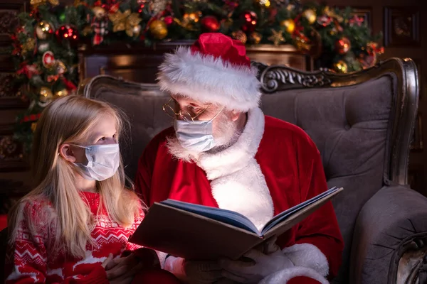 Çocuklar ve Noel Baba tıbbi maskeli. Kız ve Noel Baba evde Noel ağacının yanında oturmuş Noel kitabı okuyorlar. Mutlu yıllar, 2021.