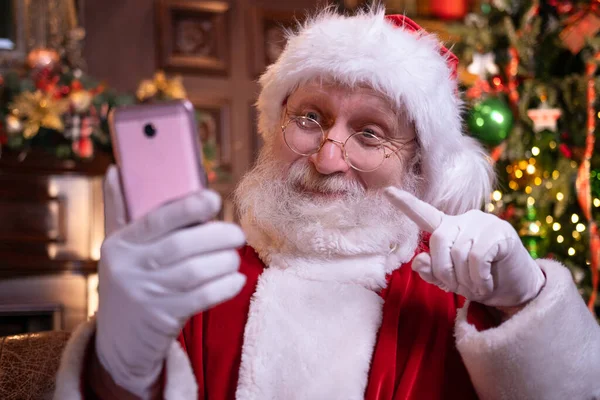 Boże Narodzenie online gratulacje od Mikołaja. Święty Mikołaj za pomocą telefonu komórkowego na odległość Christmas call ludzi i dzieci. Siedzi koło choinki w domu. Szczęśliwego Nowego Roku Covid 2021 — Zdjęcie stockowe