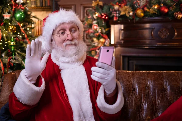 Julen online grattis från Santa. Jultomten använder mobiltelefon för distans Julsamtal människor och barn. Sitter nära julgranen hemma. Gott nytt år Covid 2021 — Stockfoto