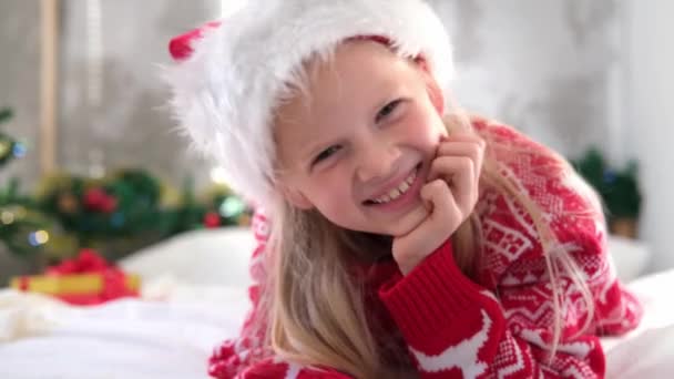 4k Πορτρέτο Χριστουγεννιάτικο κορίτσι. Χαμογελώντας παιδί στο καπέλο του Αϊ Βασίλη κοιτάζοντας στην κάμερα στο σπίτι. — Αρχείο Βίντεο