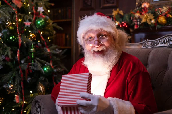 Leende jultomten håller julklapp. Uppfyllande av önskningar. Tomten sitter hemma nära julgranen — Stockfoto