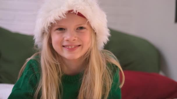 Portret świątecznej dziewczyny. Uśmiechnięte dziecko w kapeluszu Świętego Mikołaja patrzące w kamerę w domu. — Wideo stockowe