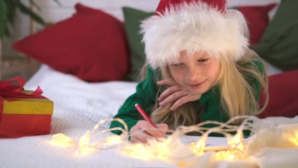 Önskelistan över julmirakel. Leende flicka i jultomten hatt skriva brev drömmar för gåvor till jultomten. Barn ligger på sängen hemma med julbelysning. — Stockvideo