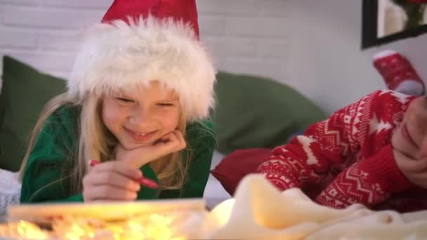 Seznam přání k Vánocům. Usmívající se chlapec a dívka v klobouku Santa Claus psaní dopis sny pro dárky Santa Claus. Děti bratr a sestra ležící v posteli se baví doma. — Stock video