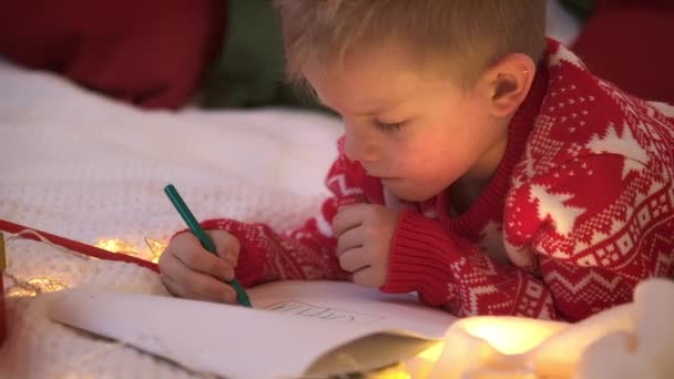 Kerstwonder verlanglijstje. Glimlachende jongen die brievendromen schrijft voor cadeaus aan de kerstman. Kind liggend op bed thuis met kerstverlichting. — Stockvideo