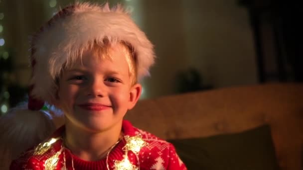 Portret uśmiechniętego chłopca w kapeluszu Mikołaja i świąteczny kostium siedzącego w domu. Nowy Rok i święta. Szczęśliwe dziecko. Spełnianie życzeń, marzeń i niespodzianek świątecznych dla dzieci. — Wideo stockowe