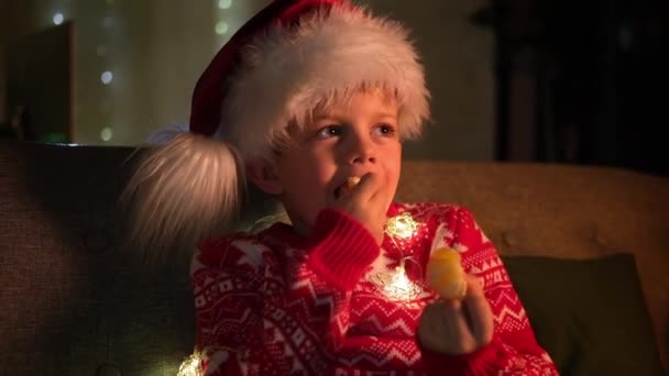 Ritratto ragazzo sognatore in cappello di Babbo Natale e costume di Natale seduto a casa, mangiare serata mandarino arancione. Capodanno e vacanze. Felicità di Natale. — Video Stock