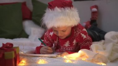 Noel mucizesi dilek listesi. Noel Baba şapkalı gülümseyen çocuk Noel Baba 'ya hediyeler için mektuplar yazıyor. Çocuk Noel ışıklarıyla evde yatıyordu..