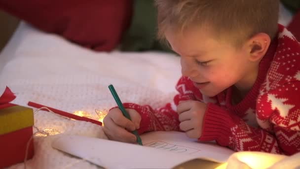 Liste de souhaits miracle de Noël. Un garçon souriant écrivant des rêves de lettre pour des cadeaux au Père Noël. Enfant couché sur le lit à la maison avec des lumières de Noël. — Video