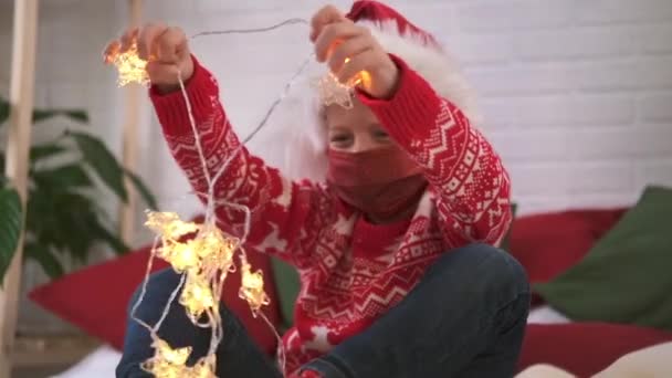 Kerst kindermasker. Jongen in de kerstman hoed versiert kamer met kerstverlichting, de voorbereiding van de vakantie thuis. — Stockvideo