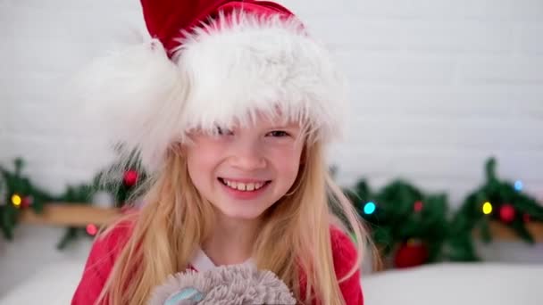 Портрет рождественской девушки. Улыбающийся ребенок в шляпе Санта-Клауса весело смотрит в камеру делает сумасшедшее смешное лицо с рождественскими яйцами на глазах — стоковое видео