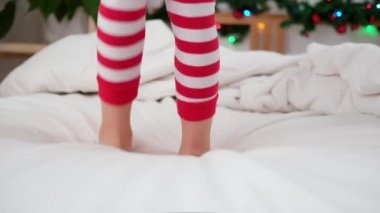 Beyaz yatakta zıplayarak bacaklarını kapat. Heyecanlı kız evde Noel pijamalarıyla dans ederken eğleniyor. Yeni yıl ve tatiller. Noel mutluluğu