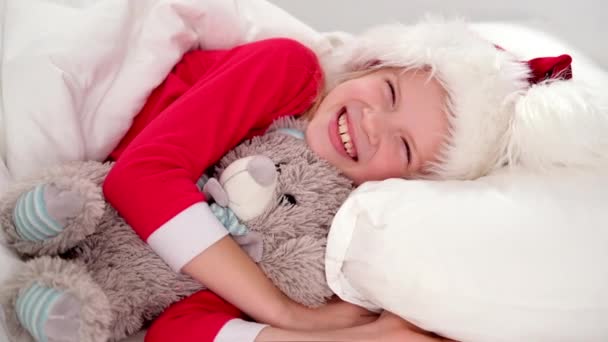 Menina sorridente em chapéu de Papai Noel e pijama de Natal deitado na cama com ursinho de pelúcia em casa. Ano Novo e Férias. Manhã de Natal felicidade. — Vídeo de Stock