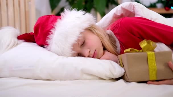 Dziewczynka w kapeluszu Mikołaja i świąteczna piżama leżąca w łóżku z pudełkiem prezentów w domu. Nowy Rok i święta. Boże Narodzenie rano szczęście. — Wideo stockowe