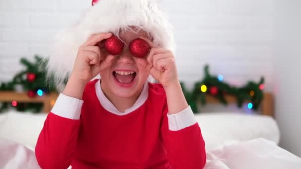 Portret bożonarodzeniowy. Uśmiechnięte dziecko w kapeluszu Świętego Mikołaja bawiące się patrząc w kamerę robiąc szalenie zabawną minę z kulkami świątecznymi na oczach — Wideo stockowe