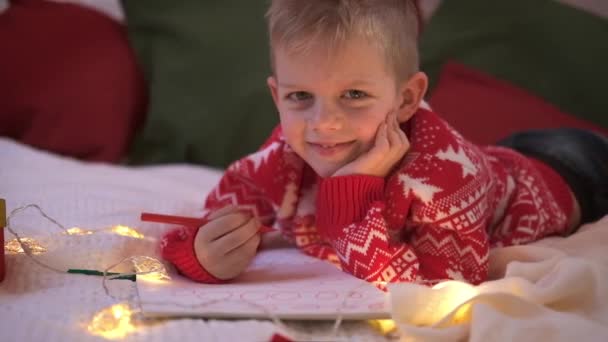 Рождественский список чудес. Улыбающийся мальчик пишет письма, мечтая о подарках Санта Клаусу. Ребенок лежит на кровати дома с рождественскими огнями. — стоковое видео