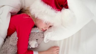 Çocuk Noel 'den önce uyuyor. Noel Baba şapkası ve Noel pijamalarıyla beyaz yatakta uzanmış oyuncak ayıyı kucaklayan küçük bir kız. Yeni yıl ve tatiller. sabah mutluluğu.