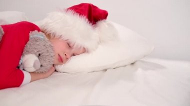 Çocuk Noel 'den önce uyuyor. Noel Baba şapkası ve Noel pijamalarıyla beyaz yatakta uzanmış oyuncak ayıyı kucaklayan küçük bir kız. Yeni yıl ve tatiller. sabah mutluluğu.