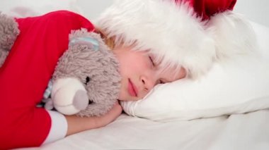 Çocuk Noel 'den önce uyuyor. Noel Baba şapkası ve Noel pijamalarıyla beyaz yatakta uzanmış oyuncak ayıyı kucaklayan küçük bir kız. Yeni yıl ve tatiller. sabah mutluluğu.