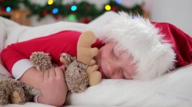 Çocuk Noel 'den önce uyuyor. Noel Baba şapkalı ve Noel pijamalı küçük çocuk beyaz yatakta uzanmış evde pelüş oyuncakları kucaklıyor. Yeni yıl ve tatiller. sabah mutluluğu.
