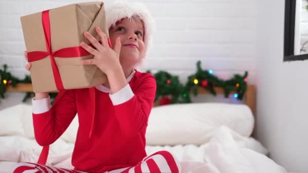 A kisfiú megrázza az ajándékdobozt, hogy megtudja, mi van benne. Karácsonyi Mikulás sapkás és pizsamás gyerek. Beteljesülő gyerekek Karácsonyi kívánságok, álmok és meglepetések — Stock videók