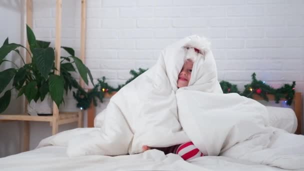 Menino de Natal animado se divertindo escondido sob as capas em chapéu de Papai Noel e pijama vermelho em casa. Ano Novo e felicidade de Natal — Vídeo de Stock