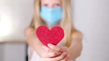 Kırmızı kalp, Sevgililer Günü 'nde tıbbi maskeli küçük sarışın kızın ellerinde. Güvenli Covid bayram kutlaması. Yavaş çekim.