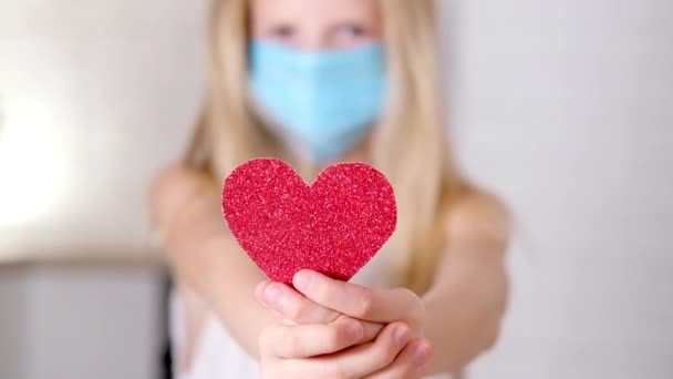 Red heart close up in hands of little blonde girl in medical mask on Valentines Day. Safety Covid holiday celebration. Slow motion. — Stock Video