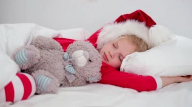 Çocuk Noel 'den önce uyuyor. Noel Baba şapkası ve Noel pijamalarıyla beyaz yatakta uzanmış oyuncak ayıyı kucaklayan küçük bir kız. Yeni yıl ve tatiller. sabah mutluluğu.