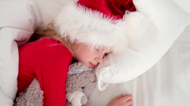 Top view Dziecko śpi przed Bożym Narodzeniem. Mała dziewczynka w kapeluszu Mikołaja i świąteczna piżama leżąca w białym łóżku przytulająca misia w domu. Nowy Rok i święta. poranne szczęście. — Wideo stockowe