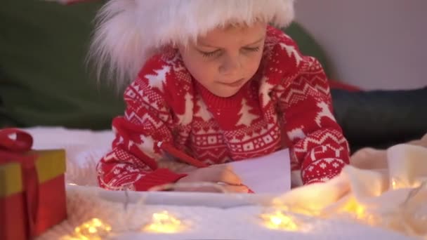 Liste de souhaits miracle de Noël. Garçon souriant dans le chapeau du Père Noël écrivant des rêves de lettre pour des cadeaux au Père Noël. Enfant couché sur le lit à la maison avec des lumières de Noël. — Video