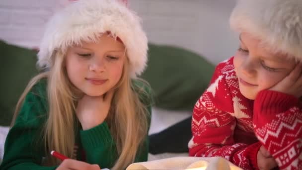 Λίστα ευχών Χριστουγέννων. Χαμογελαστό αγόρι και κορίτσι με το καπέλο του Αϊ Βασίλη που γράφει όνειρα με γράμματα για δώρα στον Αϊ Βασίλη. Παιδιά αδελφός και αδελφή ξαπλωμένοι στο κρεβάτι στο σπίτι με χριστουγεννιάτικα φώτα. — Αρχείο Βίντεο
