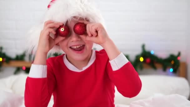 Портретный рождественский мальчик. Улыбающийся ребенок в шляпе Санта-Клауса веселится, глядя в камеру, делая сумасшедшее смешное лицо с рождественскими шарами на глазах — стоковое видео