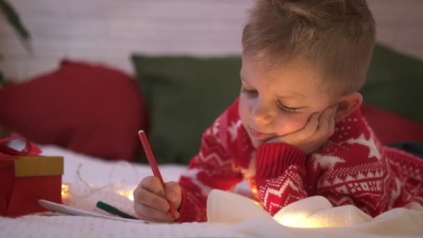 Liste de souhaits miracle de Noël. Un garçon souriant écrivant des rêves de lettre pour des cadeaux au Père Noël. Enfant couché sur le lit à la maison avec des lumières de Noël. — Video