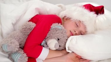 Çocuk Noel 'den önce uyuyor. Noel Baba şapkası ve Noel pijamalarıyla beyaz yatakta uzanmış oyuncak ayıyı kucaklayan küçük bir kız. Yeni yıl ve tatiller. sabah mutluluğu.